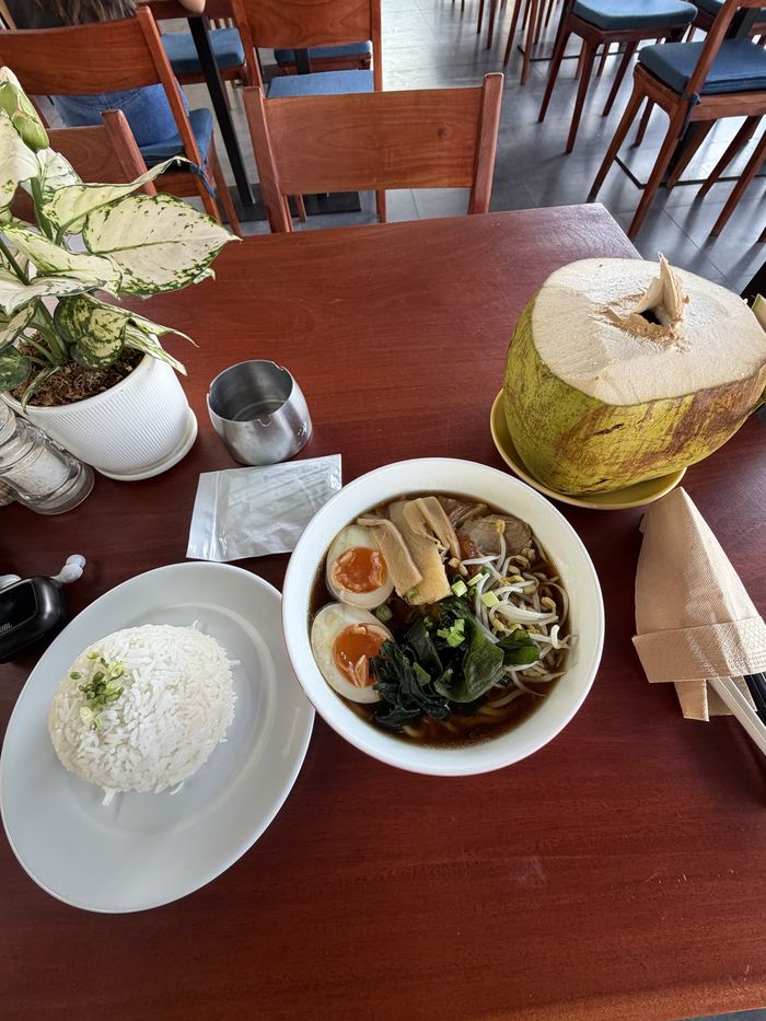 A bowl of ramen with rice and a fresh coconut on a cafe table