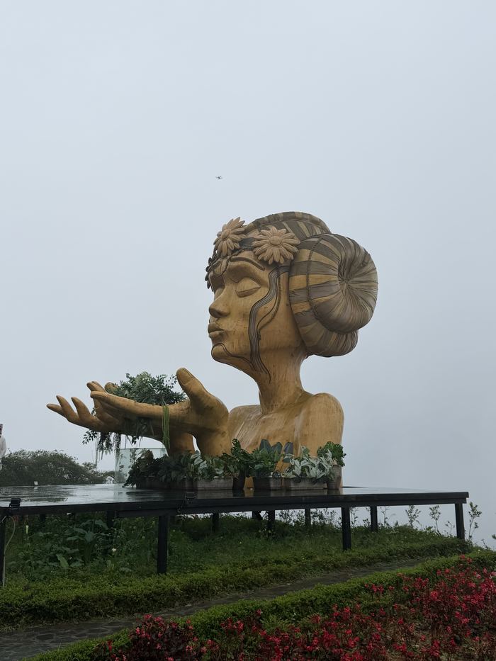 A giant sculpture of a woman's head and hands above a flower garden